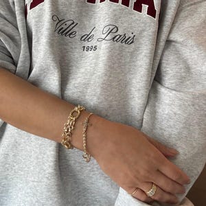 May include: Two gold bracelets are worn on an arm, one with a chain link design and the other with a cross pendant. The person is wearing a gray sweatshirt with the text "Ville de Paris 1995". Several gold rings are also visible.