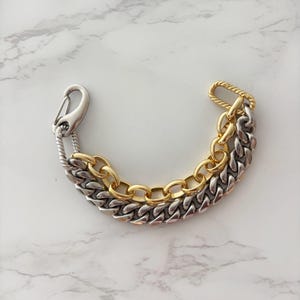 May include: A two-tone chain link bracelet featuring a mix of silver-colored and gold-colored metal links. The bracelet includes a large silver-colored lobster claw clasp and a twisted rope-textured metal bar connector at the opposite end.