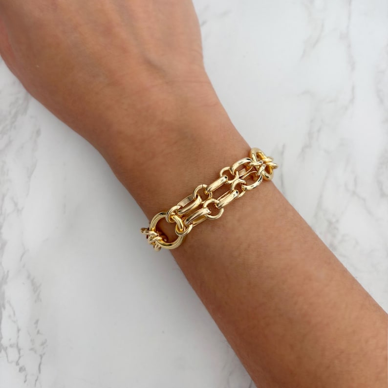 May include: A gold-colored chain link bracelet with a double-strand design. The bracelet is made of a shiny, polished metal and features a large clasp. The bracelet is shown on a person's wrist.