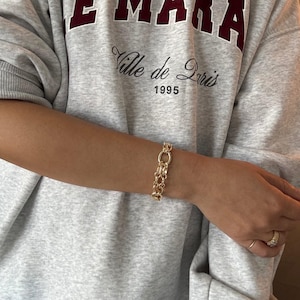 May include: A gold-colored bracelet with a chain-link design. The bracelet is worn on a person's wrist. The person is wearing a gray sweatshirt with the text "EMARA" and "Ville de Paris 1995".