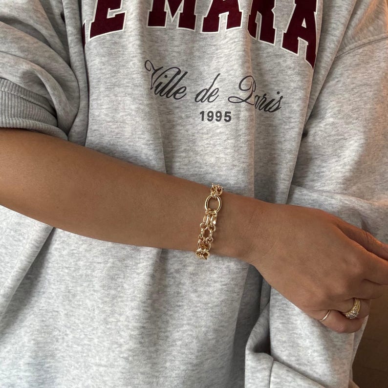 May include: A gold-colored bracelet with a chain-link design. The bracelet is worn on a person's wrist. The person is wearing a gray sweatshirt with the text "EMARA" and "Ville de Paris 1995".