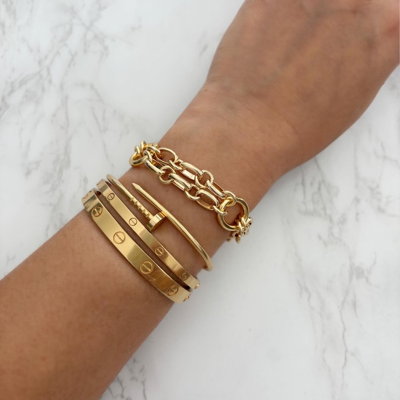 May include: A collection of gold bracelets on a wrist. The bracelets include a chain link bracelet, a nail-shaped bracelet, and several engraved bangles. The jewelry is displayed against a white and gray marble background.