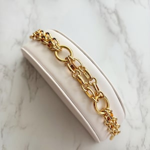 May include: A gold-toned chain bracelet with a double-strand design. The bracelet features a mix of oval and rectangular links, with large circular links connecting the strands. The jewelry is displayed on a white, angled surface.