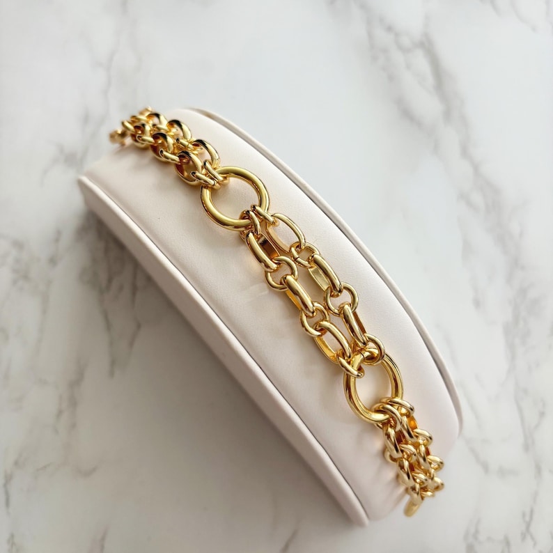 May include: A gold-toned chain bracelet with a double-strand design. The bracelet features a mix of oval and rectangular links, with large circular links connecting the strands. The jewelry is displayed on a white, angled surface.