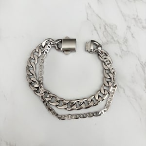 May include: A silver-colored bracelet with a double-layered chain design. The bracelet features a thick curb chain and a thinner chain, connected by a rectangular clasp. The jewelry is displayed on a white marble surface.