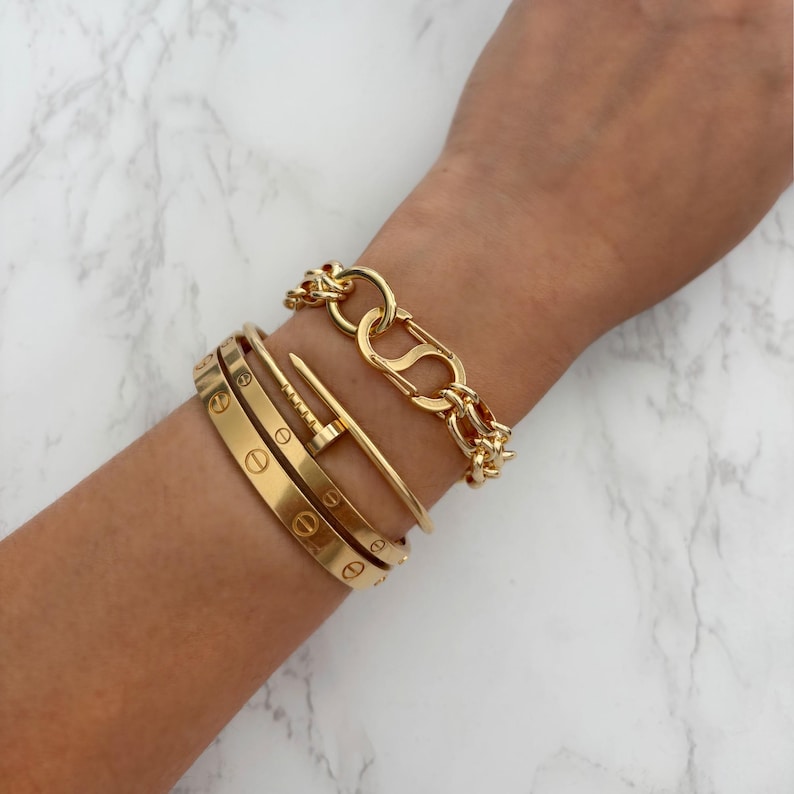 May include: A collection of gold bracelets on a wrist. The bracelets include a chain link bracelet, a nail-shaped bracelet, and two engraved bangles. The jewelry is displayed against a white and gray marble background.