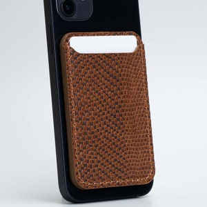May include: A brown, textured phone wallet is attached to a black smartphone. The wallet has a rectangular shape with a slot at the top for cards. The background is a plain white.