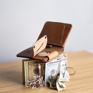 Könnte beinhalten: Ein braunes Leder-Brieftaschen-Faltmodell mit einer kleinen Münztaschenklappe, das auf einem Stapel US-Banknoten liegt, darunter Zehn- und Hundert-Dollar-Scheine, befestigt mit einer silbernen Metallklammer und einer goldenen Büroklammer.