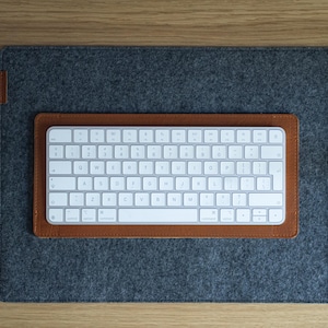 Könnte beinhalten: Eine weiße, kabellose Tastatur mit silbernen Tasten liegt auf einem braunen Lederpad, das auf einer grauen Filz-Schreibtischunterlage platziert ist. Die Tastatur hat ein Standard-QWERTZ-Layout. Das Setup befindet sich auf einer Holzoberfläche.