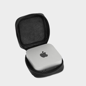 May include: A silver computer with the Apple logo is inside a black, zippered carrying case. The case is open, revealing the computer. The case is square with rounded corners and a black interior.