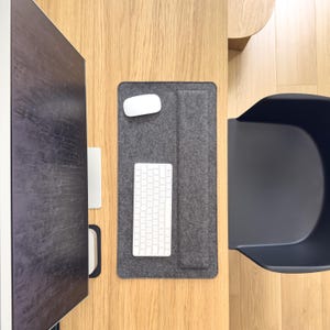 Könnte beinhalten: Draufsicht auf einen modernen Arbeitsplatz mit einer grauen Filz-Schreibtischunterlage, einer weißen kabellosen Maus und Tastatur. Ein dunkler Monitor und ein schwarzer Stuhl sind ebenfalls auf einem hellen Holzschreibtisch zu sehen.