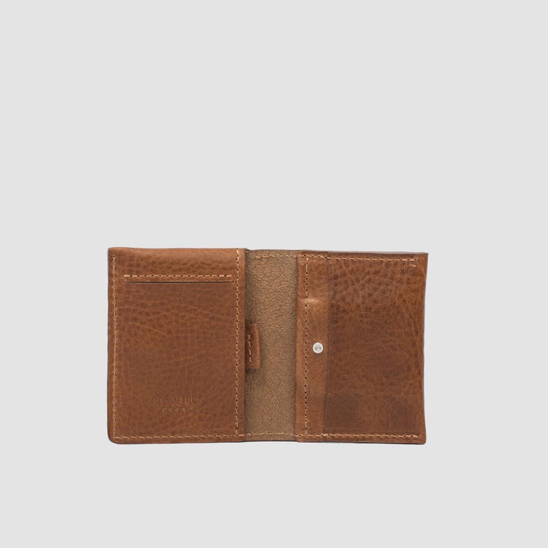 May include: A tan leather wallet featuring a textured surface, visible stitching along the edges, and a small metal snap closure on the right side. The interior displays multiple card slots and a central compartment for organizing currency and cards.