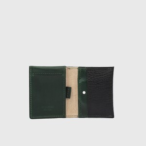 May include: A bi-fold wallet featuring a smooth dark green leather panel on the left and a textured black leather panel on the right. The interior reveals a light beige lining, a small black fabric tag, and a single silver-toned metal snap button.