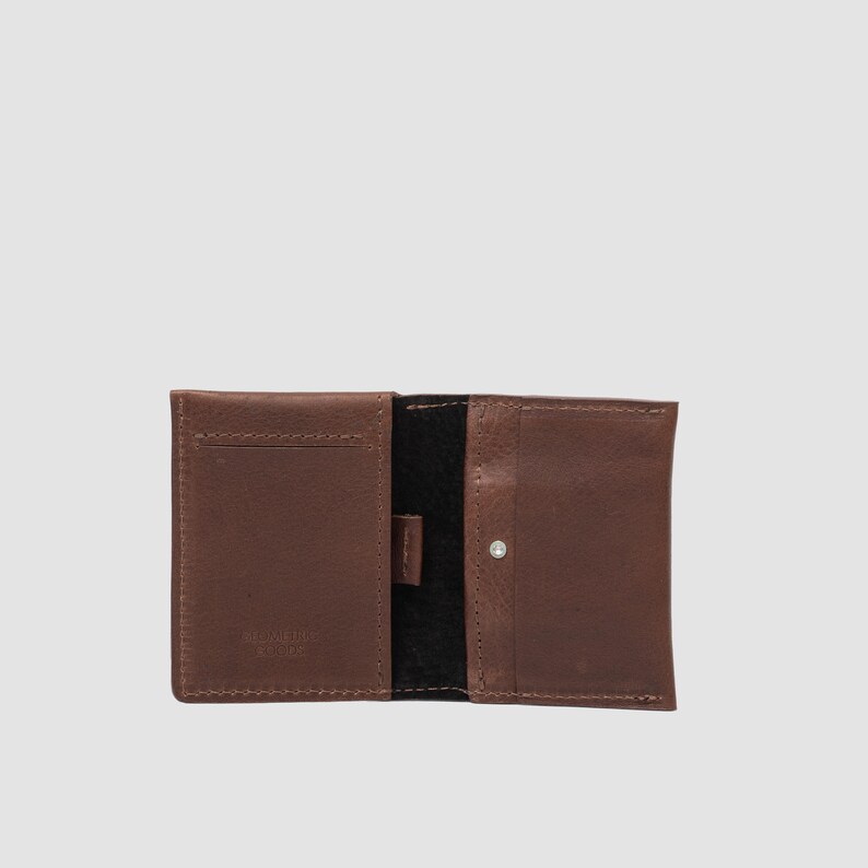 May include: A brown leather wallet featuring a bi-fold design with visible stitching along the edges. The interior displays a smooth brown leather finish on the left and right panels, with a central black suede section and a small silver metal snap.
