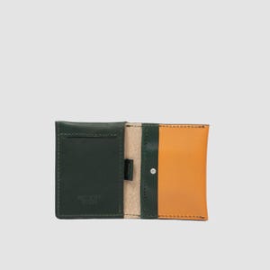 May include: A bi-fold wallet crafted from dark green and tan leather. The interior features a dark green card slot on the left, a tan leather panel on the right, and a small silver-toned metal rivet. The text "BAD STAR GOODS" is embossed on the left.