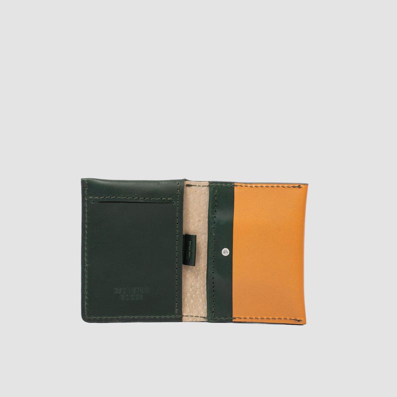 May include: A bi-fold wallet crafted from dark green and tan leather. The interior features a dark green card slot on the left, a tan leather panel on the right, and a small silver-toned metal rivet. The text "BAD STAR GOODS" is embossed on the left.