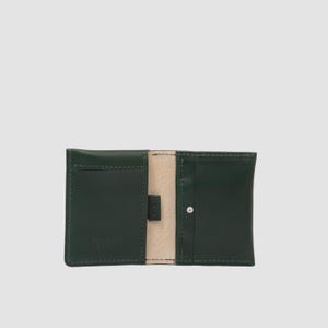 May include: A dark green leather card holder featuring a light beige interior lining. The wallet is open, displaying two main card slots on the left and right sides, with a small central tab and a single silver-toned metal rivet on the right side.