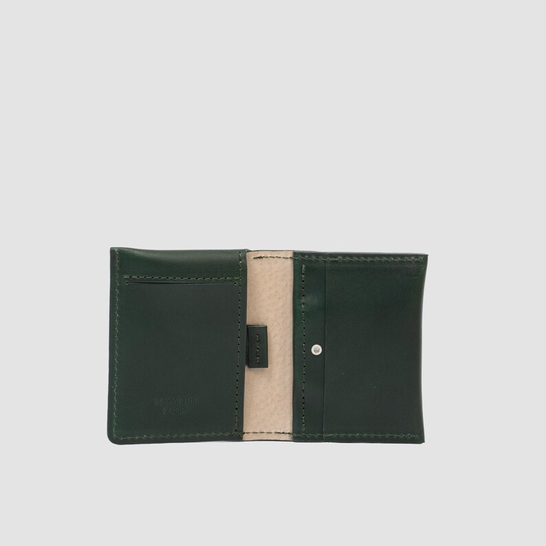 May include: A dark green leather card holder featuring a light beige interior lining. The wallet is open, displaying two main card slots on the left and right sides, with a small central tab and a single silver-toned metal rivet on the right side.