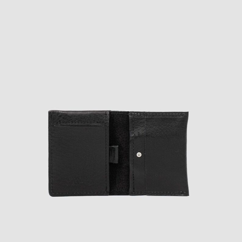 May include: A black leather wallet featuring a bi-fold design with multiple interior card slots and a central vertical strap. The exterior surface displays a textured grain, and a small silver-toned metal stud is visible on the right-hand side.