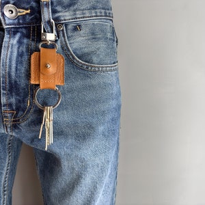 Könnte beinhalten: Ein brauner Leder-Schlüsselanhänger mit einem silbernen Karabiner und Schlüsselring, befestigt an einer blauen Jeans. Der Schlüsselanhänger hat ein rechteckiges Lederstück mit einem Druckknopfverschluss. Mehrere Schlüssel hängen an dem Ring.