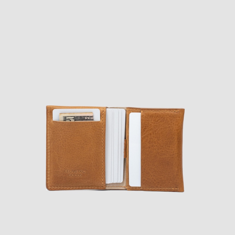 May include: A tan leather bifold wallet featuring multiple interior slots for cards and currency. The left side contains a five-dollar bill and the embossed text GEOMETRIC GOODS. The wallet is open, displaying white card edges and a smooth texture.