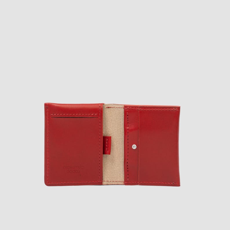 May include: A bright red leather card holder wallet shown in an open, flat position. The interior features a light beige suede lining, multiple card slots with visible stitching, and a small metal snap button closure on the right side panel.