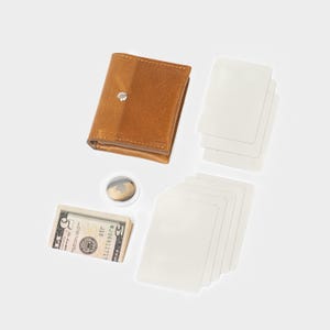 May include: A tan leather wallet with a silver snap closure, a small circular white tracking device, a stack of five-dollar bills, and several blank white rectangular cards arranged in two separate piles on a plain white background.