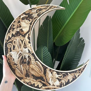 May include: A wooden crescent moon wall hanging with intricate, etched botanical and sea creature designs. The artwork features a dark brown background with light brown details, creating a striking contrast. The moon is held against a backdrop of large green leaves.