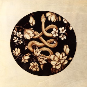 May include: A circular artwork featuring a detailed illustration of a snake intertwined with magnolia and other floral elements. The snake and flowers are in shades of brown against a black background, with a light-colored wooden surface.
