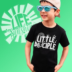 Könnte beinhalten: Ein junges Kind trägt ein schwarzes T-Shirt mit dem Text "Little Disciple" in weiß. Das Shirt zeigt einen Regenbogen, ein Kreuz und eine Krone. Das Kind trägt außerdem eine blaue Sonnenbrille und eine weiße Baseballkappe.