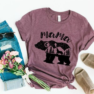 May include: A maroon t-shirt with a black silhouette of a bear with a moon, stars, and trees inside. The text "mama" is printed above the bear.