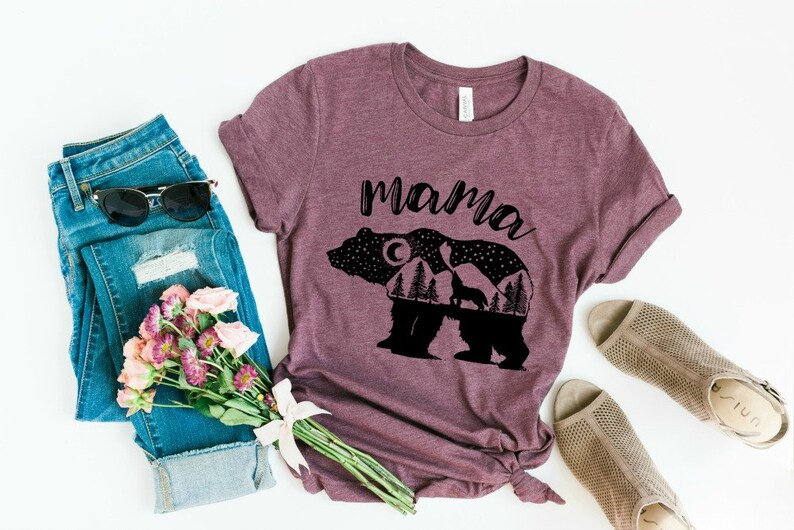 May include: A maroon t-shirt with a black silhouette of a bear with a moon, stars, and trees inside. The text "mama" is printed above the bear.
