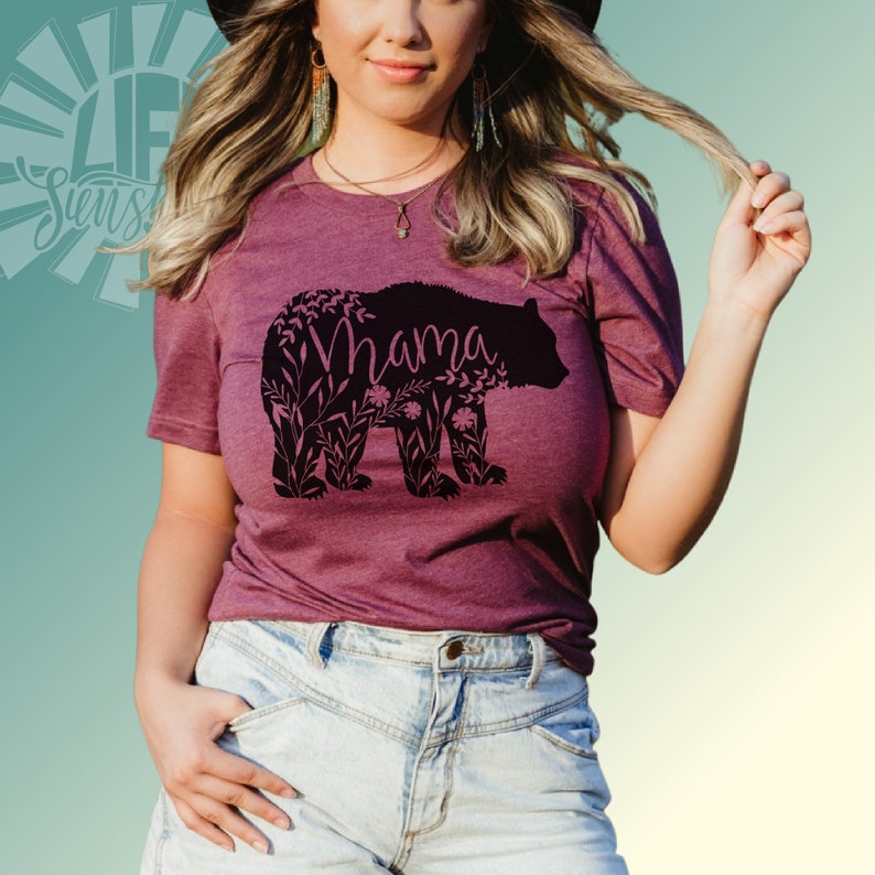 May include: A maroon t-shirt with a black silhouette of a bear with the word "Mama" inside the bear. The bear is surrounded by floral designs.