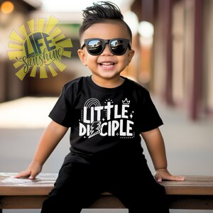 May include: A young child wearing a black t-shirt with the words "Little Disciple" in white. The shirt features a rainbow, a cross, and a crown. The child is wearing sunglasses and smiling.