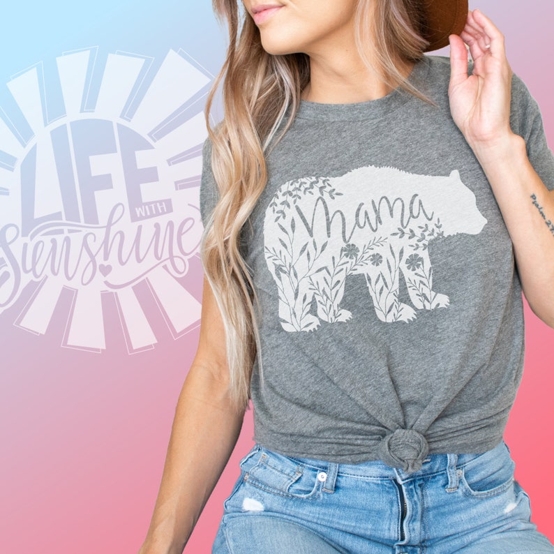 May include: Gray t-shirt with a white floral bear silhouette and the word "Mama" inside the bear. The shirt is tied at the waist and the model is wearing blue jeans.
