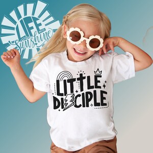 May include: White t-shirt with black text that reads "Little Disciple" with a rainbow, cross, crown, and lightning bolt graphic.