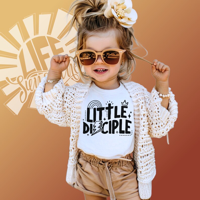 May include: A young girl wearing a white t-shirt with the text "Little Disciple" in black lettering. The shirt features a rainbow, a cross, and a crown. She is wearing a white cardigan and brown shorts.