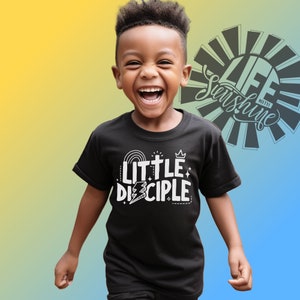 May include: A young boy wearing a black t-shirt with the text "Little Disciple" in white. The shirt features a cross, a crown, a lightning bolt, and a rainbow. The boy is smiling and looking at the camera.