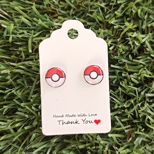 Könnte beinhalten: Ein Paar kleine, rote und weiße Pokeball-Ohrringe auf einer weißen Karte mit dem Text "Hand Made With Love Thank You" und einem roten Herzen.