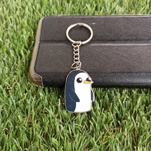 May include: A silver keyring with a cartoon penguin charm. The penguin is white with a black head and wings. It has a yellow beak and black eyes.