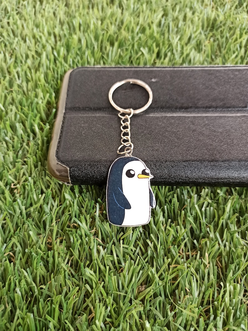May include: A silver keyring with a cartoon penguin charm. The penguin is white with a black head and wings. It has a yellow beak and black eyes.