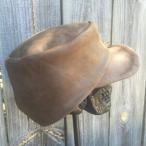 May include: A brown leather hat with a wide brim, displayed on a black metal stand. The hat has a rounded crown and a slightly curved brim. The hat is a medium brown color and appears to be made of leather.