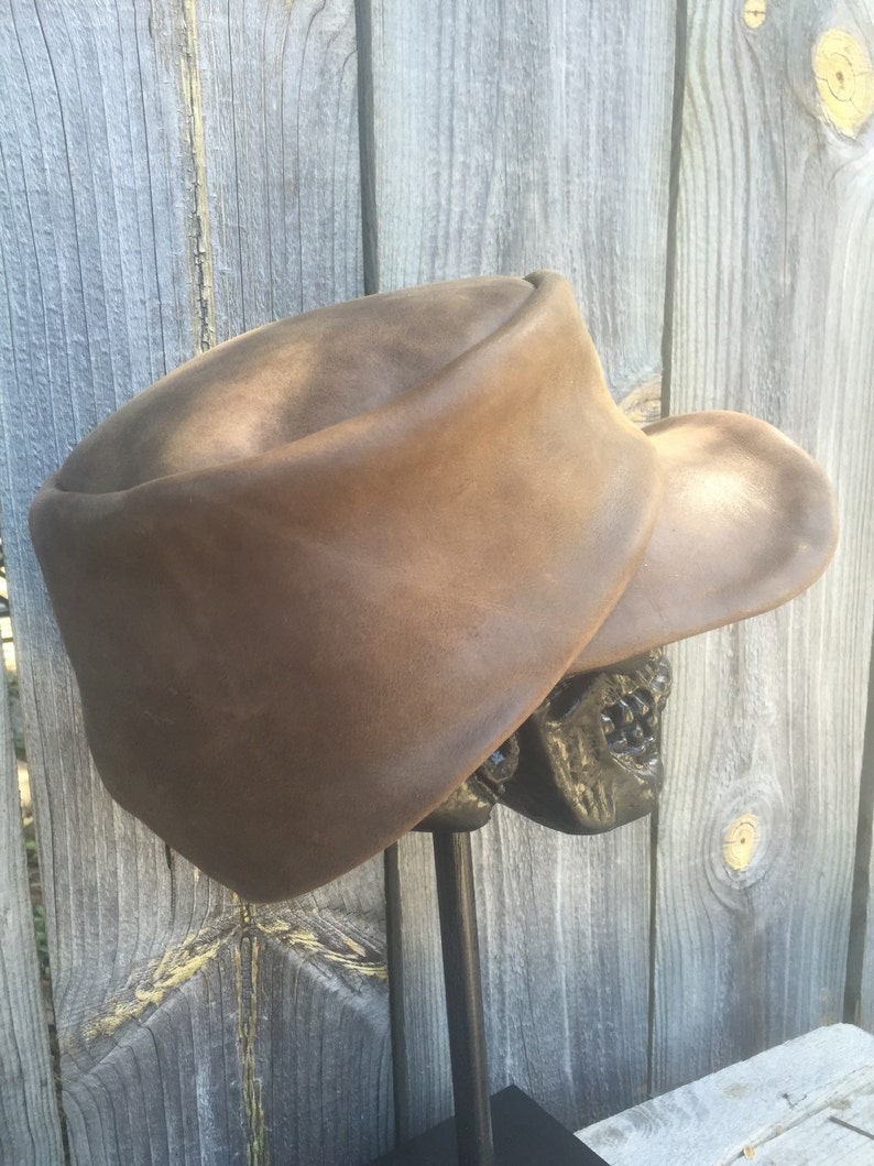 May include: A brown leather hat with a wide brim, displayed on a black metal stand. The hat has a rounded crown and a slightly curved brim. The hat is a medium brown color and appears to be made of leather.