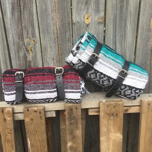 May include: Two colorful Mexican blankets with black leather straps. The blankets are rolled up and tied with the straps. The blankets are sitting on a wooden fence.