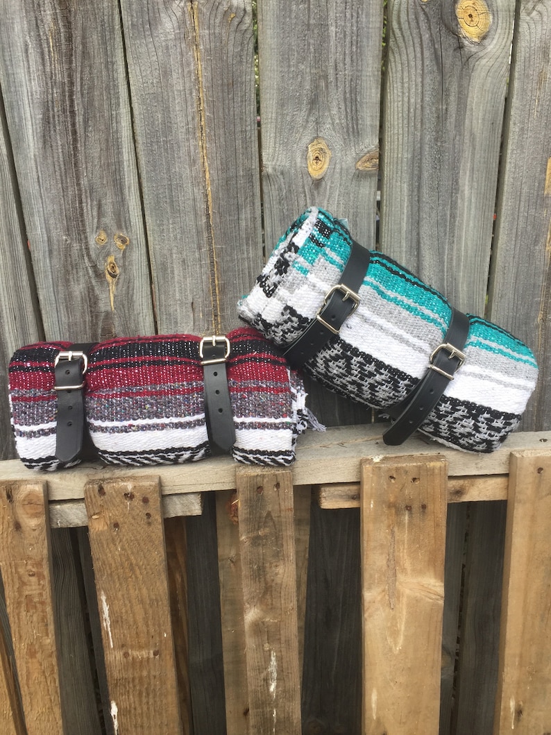 May include: Two colorful Mexican blankets with black leather straps. The blankets are rolled up and tied with the straps. The blankets are sitting on a wooden fence.
