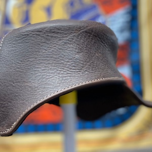 May include: A brown leather hat with a unique shape, featuring a textured surface and white stitching along the edges. The hat appears to be made of high-quality leather and is designed for style and protection.