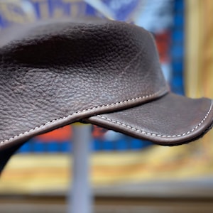 May include: A brown leather hat with a short brim. The hat has a textured surface and is stitched with white thread around the brim and seams. The hat is a dark brown color.