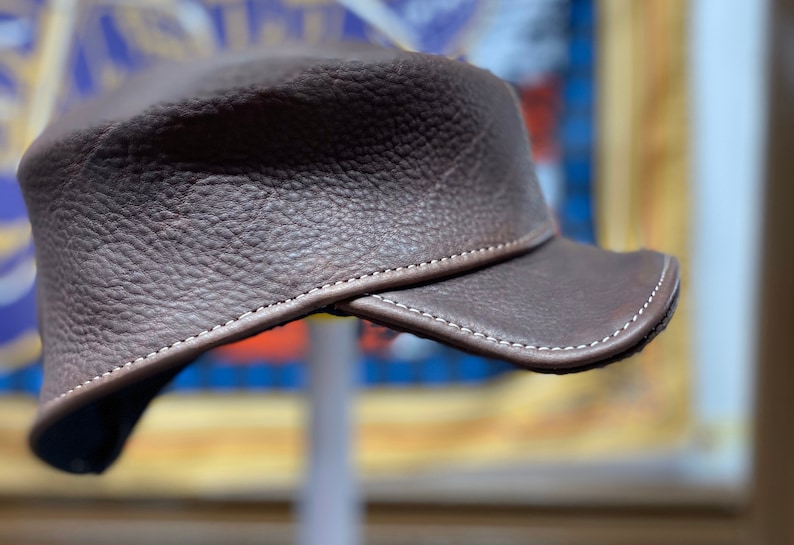 May include: A brown leather hat with a short brim. The hat has a textured surface and is stitched with white thread around the brim and seams. The hat is a dark brown color.