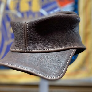 May include: A brown leather cap with a short brim. The cap features white stitching along the seams and brim. The leather has a textured appearance. The cap is a classic style, suitable for various occasions.