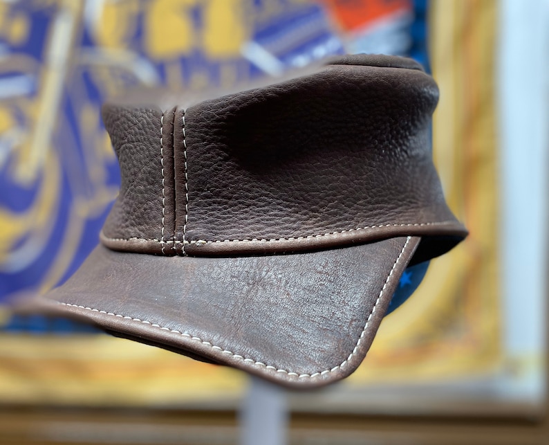 May include: A brown leather cap with a short brim. The cap features white stitching along the seams and brim. The leather has a textured appearance. The cap is a classic style, suitable for various occasions.